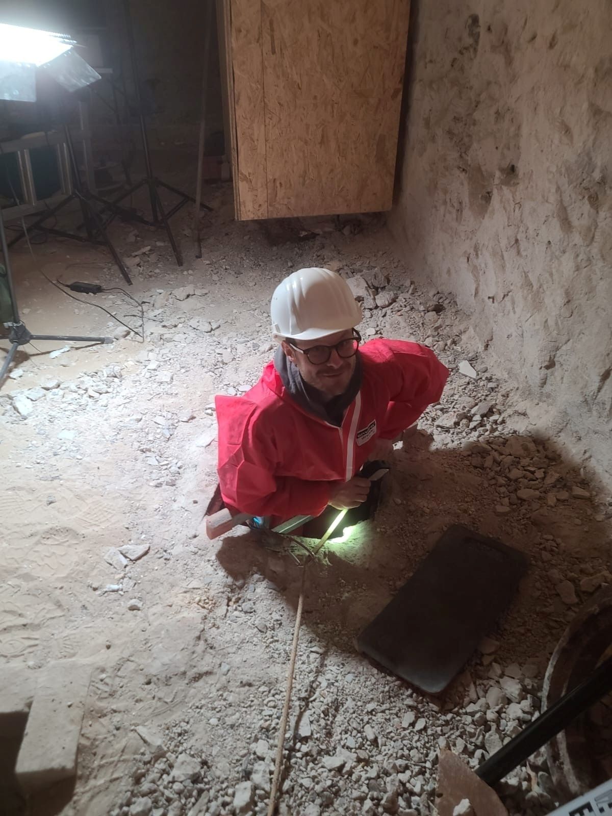 Eine Person in rotem Schutzanzug und weißem Helm steigt in ein Bodenloch in einem staubigen, unebenen Raum mit grobem Boden. Die Person hält eine Taschenlampe, die auf den Boden gerichtet ist, und befindet sich neben einer rechteckigen, dunklen Bodenplatte. Im Hintergrund steht eine Holzplatte an der Wand, und links sind Studiobeleuchtung und Kabel sichtbar. Die Wände sind rau und unregelmäßig, vermutlich aus Stein oder Beton.