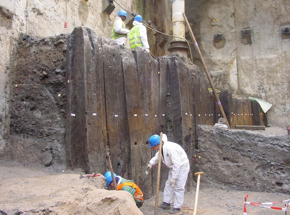 Mehrere Personen mit Schutzkleidung und blauen Helmen arbeiten an einer Ausgrabungsstätte. Im Hintergrund steht eine hohe, senkrechte Holzwand, die von Erde und Gestein umgeben ist. Die Arbeiter führen verschiedene Tätigkeiten aus, einige stehen auf der Holzkonstruktion, andere graben im Boden davor. Die Szene spielt in einer tiefen Baugrube mit sichtbaren Erdschichten.