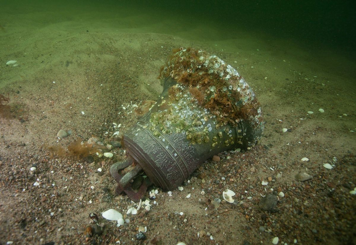 Eine große Schiffsglocke liegt halb im Sand auf dem Meeresboden. Die Oberfläche ist mit Muscheln, Algen und anderen Meeresorganismen bewachsen. Das Wasser ist grünlich und trüb, der Untergrund besteht aus Sand und kleinen Muschelstücken. Die Glocke zeigt Verzierungen und erhabene Schriftzüge, die jedoch teilweise von Bewuchs verdeckt sind.