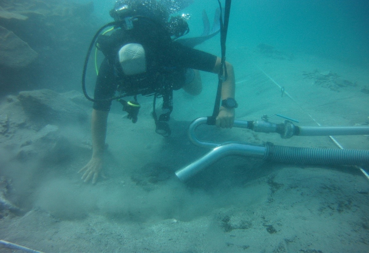 Ein Taucher in kompletter Tauchausrüstung arbeitet unter Wasser am Meeresboden. Mit einer Hand hält er einen großen Schlauch, der Sediment absaugt und dabei eine Staubwolke aufwirbelt. Im Hintergrund sind Felsen und Messlinien zu erkennen, die auf eine archäologische Ausgrabung hindeuten.