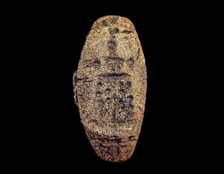 Ein ovales Siegel mit rauer, goldbrauner Oberfläche liegt vor schwarzem Hintergrund. In der Mitte sind eingravierte Linien und Muster zu erkennen, die eine stilisierte Struktur mit mehreren Ebenen und einer markanten Spitze zeigen. Die Gravuren wirken abgegriffen und sind teilweise schwer zu erkennen.