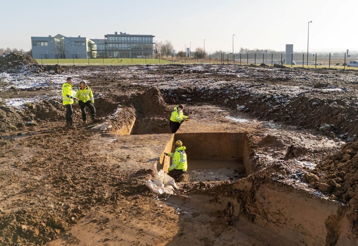 Vier Personen in gelben Warnwesten stehen und hocken in einer rechteckigen Ausgrabungsgrube auf einer schlammigen Baustelle. Die Grube ist von Erdreich umgeben, im Hintergrund sind moderne Gebäude und ein Zaun zu sehen. Der Himmel ist klar, das Gelände wirkt weitläufig.