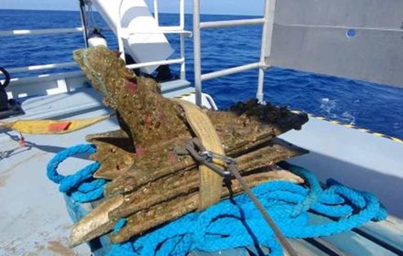 Ein Stück antiker Schiffswrackteile, mit Algen und Muscheln bedeckt, liegt auf dicken, blauen Schiffstauen an Bord eines Schifes.